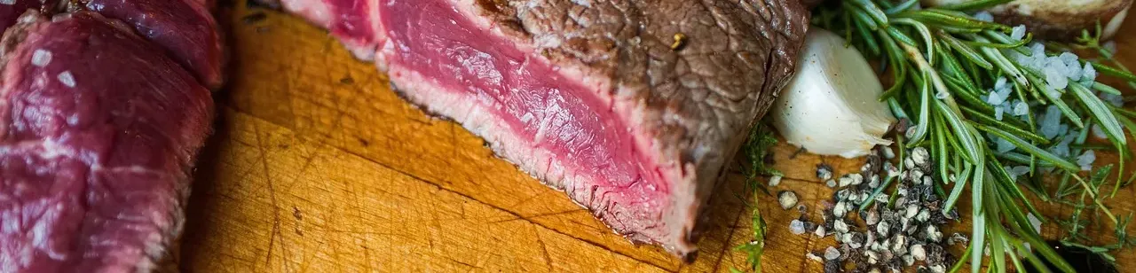 Angeschnittenes Rindersteak mit rosa-rotem Kern auf einem Holzbrett, daneben Rosmarin, Knoblauch und Pfeffer